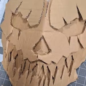 May include: A cardboard mask with a skull design. The mask has a jagged, sharp-toothed mouth and a detailed skull face.