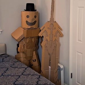 May include: A cardboard figure wearing a black top hat and holding a cardboard sword. The figure has a smiling face drawn on it. The figure is standing in a bedroom next to a bed with a patterned comforter.