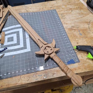 May include: A handcrafted cardboard sword with intricate detailing. The sword has a star-shaped crossguard, a circular emblem, and a wrapped handle. The blade is long and pointed, and the overall colour is brown.