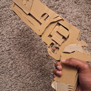May include: A handmade cardboard toy gun, featuring a detailed design with a barrel, handle, and trigger. The toy is constructed from corrugated cardboard, showcasing the material's natural brown color. The gun is held by a person.