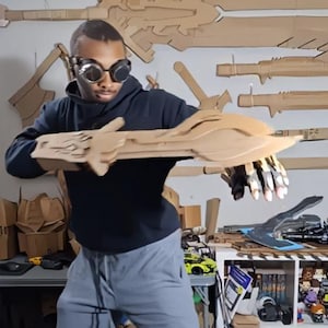 Op de afbeelding: Een persoon met een bril die een kartonnen armrekwisiet vasthoudt. De rekwisiet is gemaakt van gelaagd karton en heeft een gedetailleerd ontwerp. Kartonnen wapens zijn op de achtergrond aan de muur bevestigd.