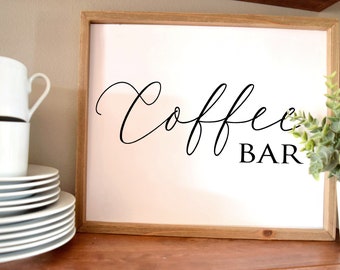 May include: A framed sign with the words "Coffee BAR" in black script. The sign has a white background and a light brown wooden frame. White coffee mugs and plates are stacked on the left side of the frame.