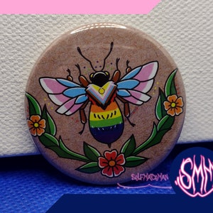 Puede incluir: Un botón redondo con un diseño de abeja colorido. La abeja tiene un abdomen rayado arcoíris y alas translúcidas con acentos rosa, azul y blanco. La abeja está rodeada de hojas verdes y flores con pétalos amarillos, naranjas y rojos.