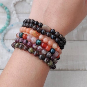 May include: A collection of beaded bracelets in various colors, including black, orange, pink, green, and purple. The bracelets are made of small, round beads and are stacked on a wrist. Other bracelets are visible in the background.