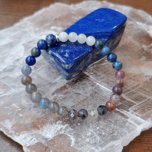 May include: A beaded bracelet featuring a variety of gemstones in shades of blue, gray, and pink. The bracelet is displayed on a white, crystalline surface, with a dark blue lapis lazuli stone in the background.