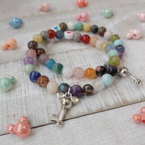 May include: A colorful beaded bracelet with a silver key charm and a variety of gemstone beads in shades of blue, green, pink, purple, and brown. The bracelet is on a white wooden surface, surrounded by decorative beads.