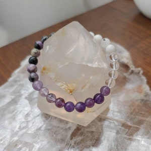 May include: A beaded bracelet featuring a gradient of purple, clear, white, and gray gemstones. The bracelet is displayed on a clear quartz crystal, showcasing the natural colors and textures of the stones.