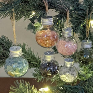 May include: Clear glass Christmas ornaments filled with colorful stones, hanging from a Christmas tree. Each ornament has a silver cap and is suspended by twine. The stones are various colors, including pink, green, and blue.