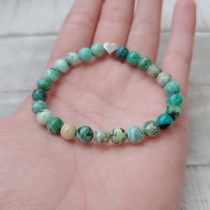 Puede incluir: Pulsera de cuentas redondas turquesas y verdes. Un peque&ntilde;o dije de coraz&oacute;n plateado est&aacute; centrado en la pulsera. La pulsera se muestra en la mano. Las cuentas miden aproximadamente 0,8 cm de di&aacute;metro.