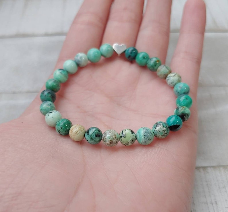 Puede incluir: Pulsera de cuentas redondas turquesas y verdes. Un peque&ntilde;o dije de coraz&oacute;n plateado est&aacute; centrado en la pulsera. La pulsera se muestra en la mano. Las cuentas miden aproximadamente 0,8 cm de di&aacute;metro.