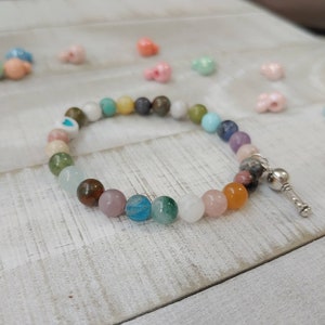 May include: A beaded bracelet featuring a variety of colorful gemstone beads. The bracelet includes a mix of colors such as pink, blue, green, and orange. A silver-colored charm is attached to the end of the bracelet. The bracelet is on a white wooden surface.