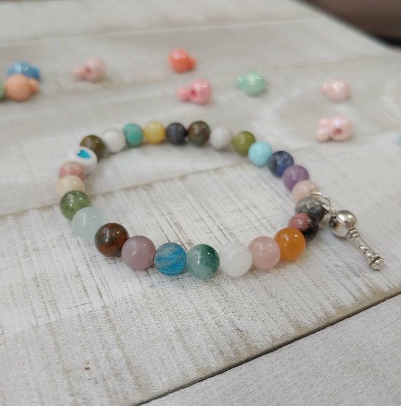 May include: A beaded bracelet featuring a variety of colorful gemstone beads. The bracelet includes a mix of colors such as pink, blue, green, and orange. A silver-colored charm is attached to the end of the bracelet. The bracelet is on a white wooden surface.