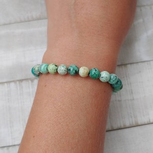 Puede incluir: Un brazalete de cuentas redondas de color turquesa con motas verde oscuro y crema. El brazalete se lleva en la mu&ntilde;eca, mostrando las cuentas de piedra natural. Es una pieza de joyer&iacute;a.