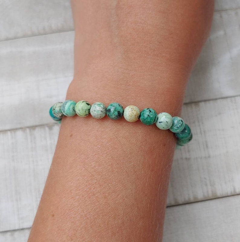 Puede incluir: Un brazalete de cuentas redondas de color turquesa con motas verde oscuro y crema. El brazalete se lleva en la mu&ntilde;eca, mostrando las cuentas de piedra natural. Es una pieza de joyer&iacute;a.