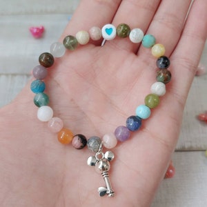May include: A beaded bracelet featuring a variety of colorful gemstone beads. The bracelet includes a charm of a silver-colored key with a Mickey Mouse head design. The beads are in shades of blue, green, pink, and orange.