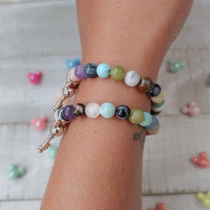 May include: Two beaded bracelets with a variety of colorful gemstones. The bracelets are silver and have a key-shaped charm. The gemstones include purple, blue, green, and white beads.