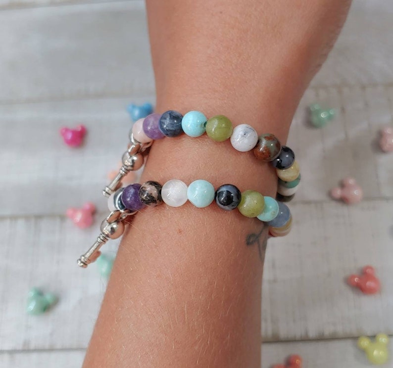 May include: Two beaded bracelets with a variety of colorful gemstones. The bracelets are silver and have a key-shaped charm. The gemstones include purple, blue, green, and white beads.
