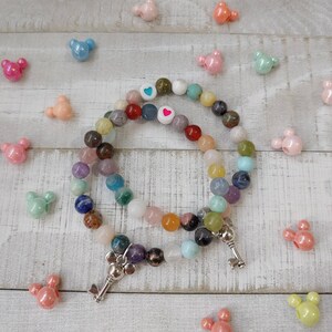 May include: Two beaded bracelets with a variety of colorful gemstones and silver key charms. The bracelets are surrounded by Mickey Mouse-shaped beads in various pastel colors. The bracelets have white beads with heart and blue heart designs.