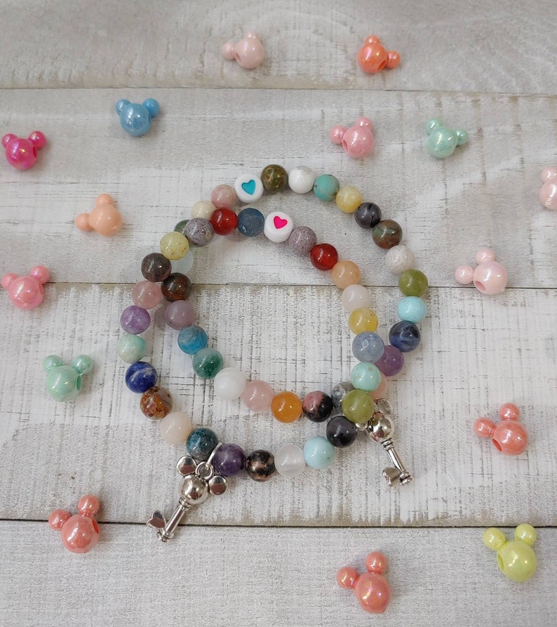 May include: Two beaded bracelets with a variety of colorful gemstones and silver key charms. The bracelets are surrounded by Mickey Mouse-shaped beads in various pastel colors. The bracelets have white beads with heart and blue heart designs.