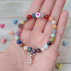 May include: A beaded bracelet with a variety of colorful gemstone beads. The bracelet features a white bead with a pink heart and a silver key charm. The beads are in shades of red, blue, green, and pink.