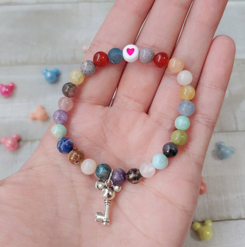 May include: A beaded bracelet with a variety of colorful gemstone beads. The bracelet features a white bead with a pink heart and a silver key charm. The beads are in shades of red, blue, green, and pink.