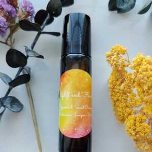 May include: A black essential oil roller bottle with a colorful label that reads "Uplift and Focus." The label features a watercolor design with yellow, orange, and pink hues. The bottle is surrounded by dried flowers and greenery.