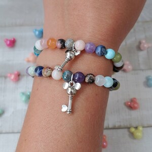 May include: Two beaded bracelets with a variety of colorful gemstones and silver key charms. The bracelets are composed of round beads in shades of pink, purple, blue, green, and brown. The key charms have a Mickey Mouse head design.