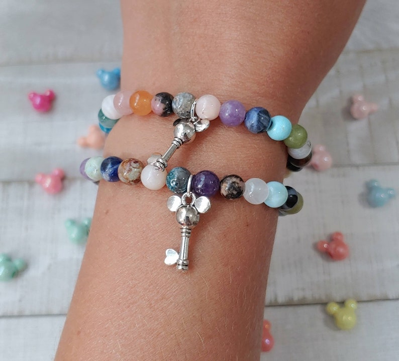 May include: Two beaded bracelets with a variety of colorful gemstones and silver key charms. The bracelets are composed of round beads in shades of pink, purple, blue, green, and brown. The key charms have a Mickey Mouse head design.