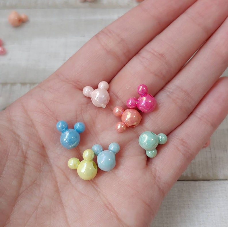 May include: A collection of colorful, pearlescent Mickey Mouse-shaped beads in various pastel shades, including pink, blue, yellow, and green. The beads are small and round, with a glossy finish, perfect for crafting projects.