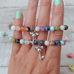 May include: Two beaded bracelets with a variety of colorful gemstones. Each bracelet features a silver key charm. The gemstones include white, pink, blue, green, and brown beads. The bracelets are displayed on a hand.