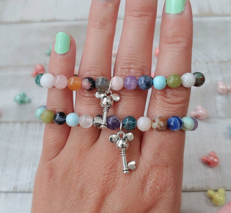 May include: Two beaded bracelets with a variety of colorful gemstones. Each bracelet features a silver key charm. The gemstones include white, pink, blue, green, and brown beads. The bracelets are displayed on a hand.