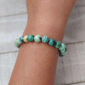 Puede incluir: Una pulsera de cuentas redondas de color turquesa, con vetas verdes oscuras y blancas. Las cuentas est&aacute;n ensartadas, creando una joya. La pulsera se lleva en un brazo, destacando los patrones y colores de la piedra natural.