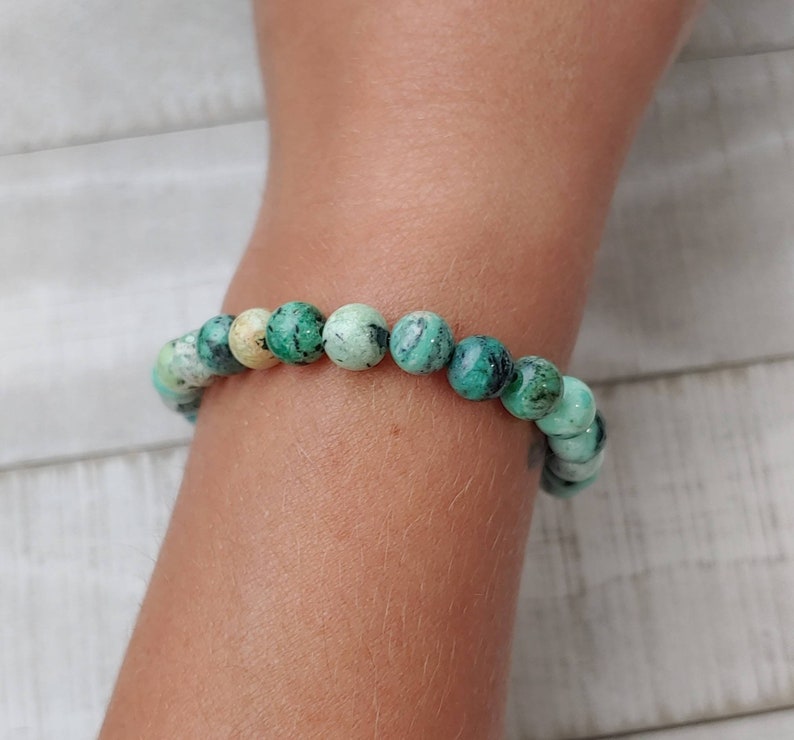 Puede incluir: Una pulsera de cuentas redondas de color turquesa, con vetas verdes oscuras y blancas. Las cuentas est&aacute;n ensartadas, creando una joya. La pulsera se lleva en un brazo, destacando los patrones y colores de la piedra natural.