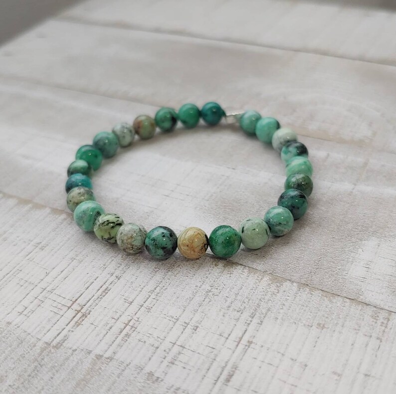 Puede incluir: Un brazalete de cuentas redondas de color turquesa, con variaciones en tonos verdes y beige. El brazalete est&aacute; ensartado en un cord&oacute;n el&aacute;stico transparente y descansa sobre una superficie de madera clara.