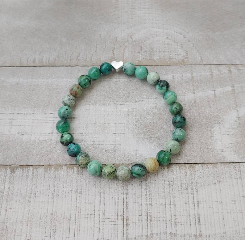 Puede incluir: Pulsera de cuentas verdes y turquesas con un peque&ntilde;o dije de coraz&oacute;n plateado. Las cuentas redondas son de piedra natural. La pulsera es el&aacute;stica.