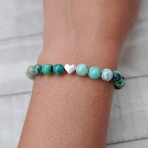 Puede incluir: Una pulsera de cuentas redondas turquesas y negras moteadas. Un peque&ntilde;o dije de coraz&oacute;n plateado est&aacute; centrado en la pulsera. La pulsera se lleva en la mu&ntilde;eca.