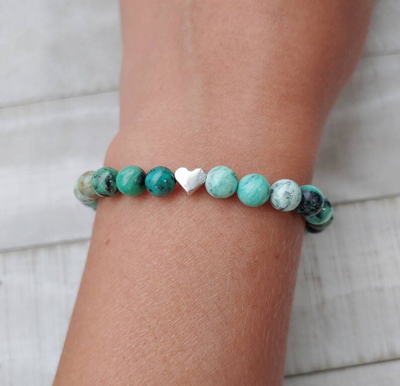 Puede incluir: Una pulsera de cuentas redondas turquesas y negras moteadas. Un peque&ntilde;o dije de coraz&oacute;n plateado est&aacute; centrado en la pulsera. La pulsera se lleva en la mu&ntilde;eca.