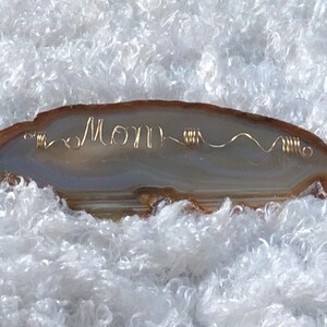 May include: A light brown agate slice with a gold wire wrapped around it, spelling out the word "Mom".