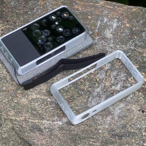 May include: A rectangular digital camera with multiple lenses and a black screen, resting on a gray pad. A clear, rectangular frame and a black strap are nearby. The camera has the word "light" printed on it.