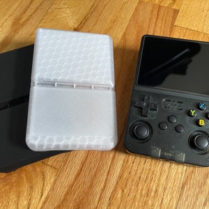 May include: A black handheld gaming console with a screen, buttons, and a joystick. The console is next to a white protective case with a honeycomb pattern.