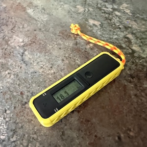 May include: A yellow and black digital device with a screen displaying "18.1". The device has a textured yellow casing and a black top with a button. An orange and yellow lanyard is attached to the device.