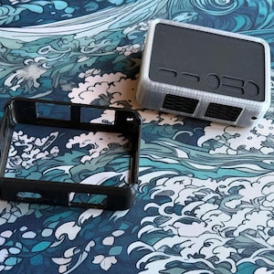 May include: A black and gray electronic device with a matching black frame, set against a blue and white wave pattern. The device has a rectangular shape with vents and buttons. The frame is open, revealing the device's interior.