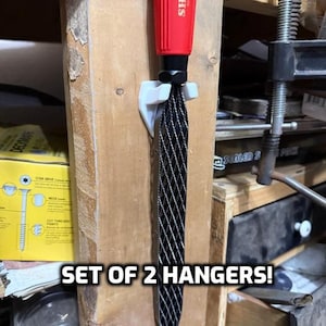 May include: A set of two white plastic tool hangers with a black metal rasp hanging on one of them. The text "SET OF 2 HANGERS!" is visible in the image.