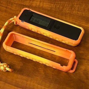 May include: A yellow and orange protective case for a small electronic device with a digital display showing the number 3.96. The case has a lanyard attached.