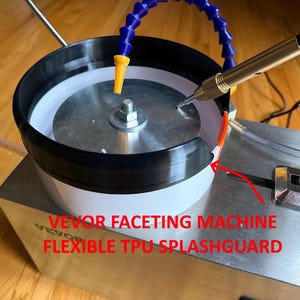 May include: A faceting machine with a black flexible TPU splashguard. The machine has a silver metal base and a circular grinding area. A blue and yellow flexible tube is positioned above the grinding area. The text "VEVOR FACETING MACHINE FLEXIBLE TPU SPLASHGUARD" is visible.