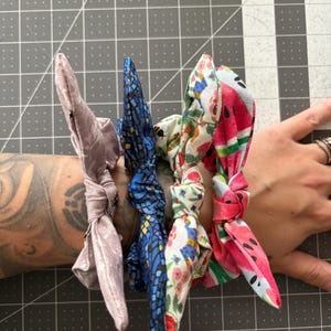 May include: Four fabric scrunchies in various patterns and colors. One is a light purple floral print, another is blue with a mosaic design, a third has a floral pattern, and the last is a watermelon print. The scrunchies are tied in a bow.