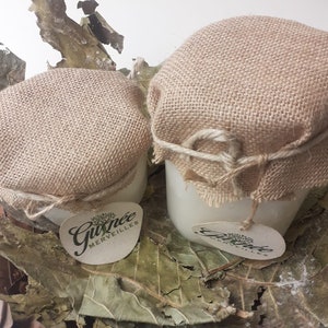 Puede incluir: Dos tarros blancos con tapas de arpillera atadas con hilo, cada uno con una pequeña etiqueta que dice "Gusnée Merveilles" en escritura verde. Los tarros descansan sobre un lecho de hojas secas.