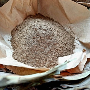 May include: A pile of brown powder sits in a crumpled brown paper bag. The powder is finely ground and has a uniform texture.