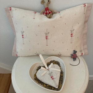 Könnte beinhalten: Ein weißes Kissen mit einem rosa-roten gestreiften Rand zeigt ein sich wiederholendes Muster aus grauen Mäusen, die rote Tutus tragen. Eine kleine graue Maus, die ein rot-weiß kariertes Kleid trägt, sitzt auf dem Kissen. Ein weißes herzförmiges Kissen, gefüllt mit getrockneter Lavendel, steht auf einem weißen Tisch.