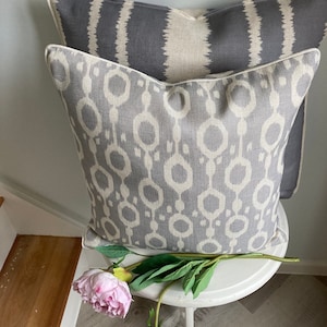 May include: Two decorative throw pillows with a geometric pattern in shades of grey and white. The pillows are sitting on a white stool with a pink flower.